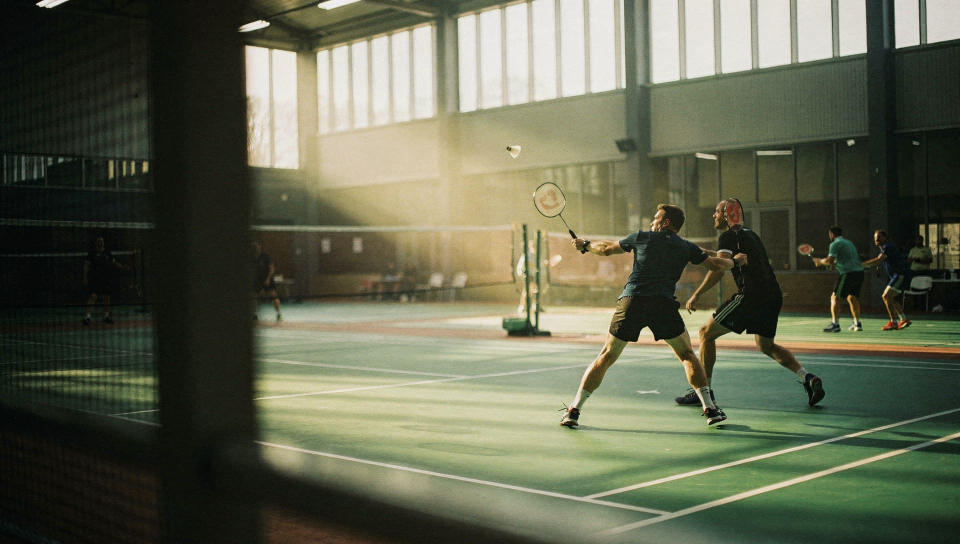 A doubles rally in a sunlit badminton hall